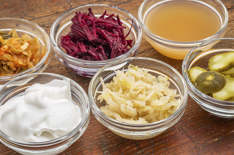 a sampler of fermented food great for gut health - glass bowls against wood: kimchi, red beets, apple cider vinegar, coconut milk yogurt, cucumber pickles, sauerkraut